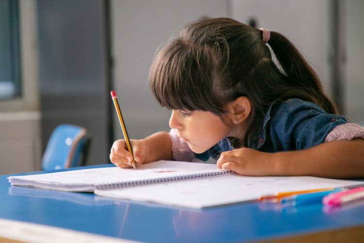 Focused black haired Latin girl sitting at school desk and drawing in her copybook. Education or back to school concept