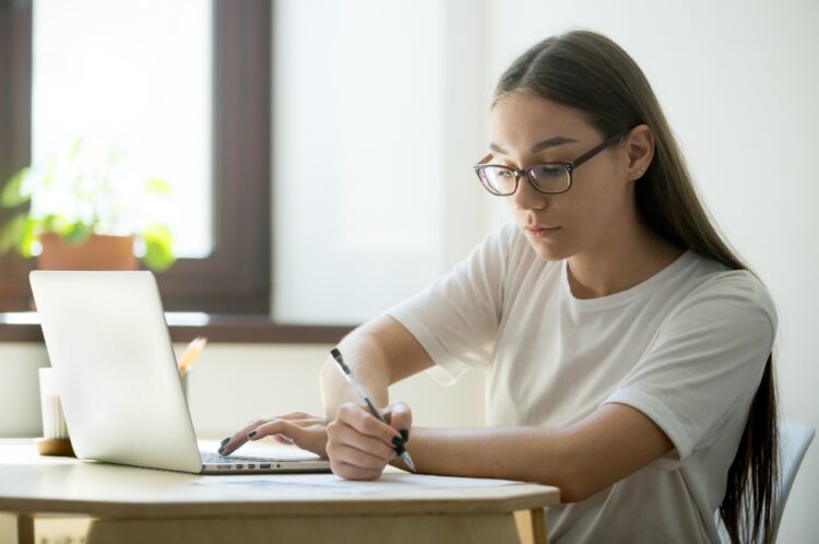 Concentrated serious student working at laptop, studying, writing down important information, processing data. Intern training online, taking web courses