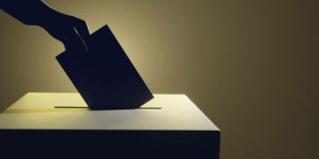 Silhouette of a hand putting a vote into the voting box on pale yellow background. Illustration of the concept of legislative election