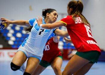 Two athletes in a handball match compete for the ball, one in a blue and white uniform and the other in red and green, with spectators visible in the background.
