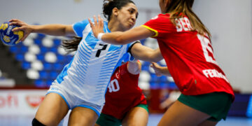 Two athletes in a handball match compete for the ball, one in a blue and white uniform and the other in red and green, with spectators visible in the background.