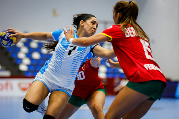 Dura caída de La Garrita en su debut en el Mundial U18 Two athletes in a handball match compete for the ball, one in a blue and white uniform and the other in red and green, with spectators visible in the background.