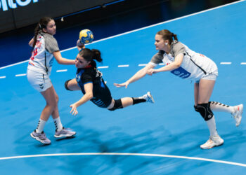 Three athletes compete intensely in a handball match, with one player diving to catch the ball while two opponents attempt to block her.