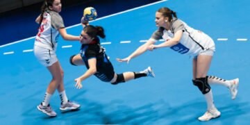 Three athletes compete intensely in a handball match, with one player diving to catch the ball while two opponents attempt to block her.