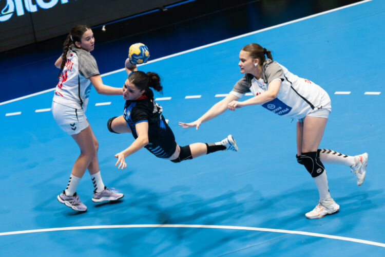 La Garrita no consolidó la levantada: perdió con Serbia y peleará por el 23º puesto Three athletes compete intensely in a handball match, with one player diving to catch the ball while two opponents attempt to block her.
