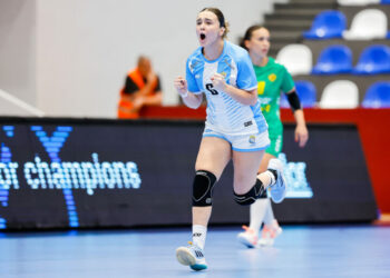 An athlete in a light blue and white uniform celebrates with a raised fist during an indoor handball game.