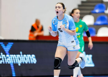 An athlete in a light blue and white uniform celebrates with a raised fist during an indoor handball game.