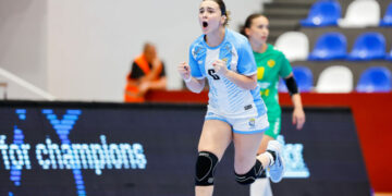 An athlete in a light blue and white uniform celebrates with a raised fist during an indoor handball game.